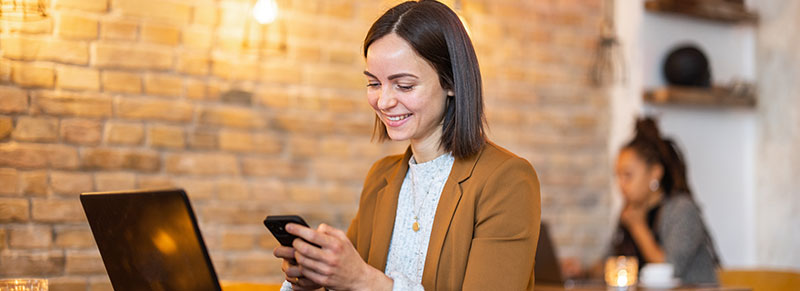 mujer sentada frente a la computadora, sonrie mientras revisa su celular